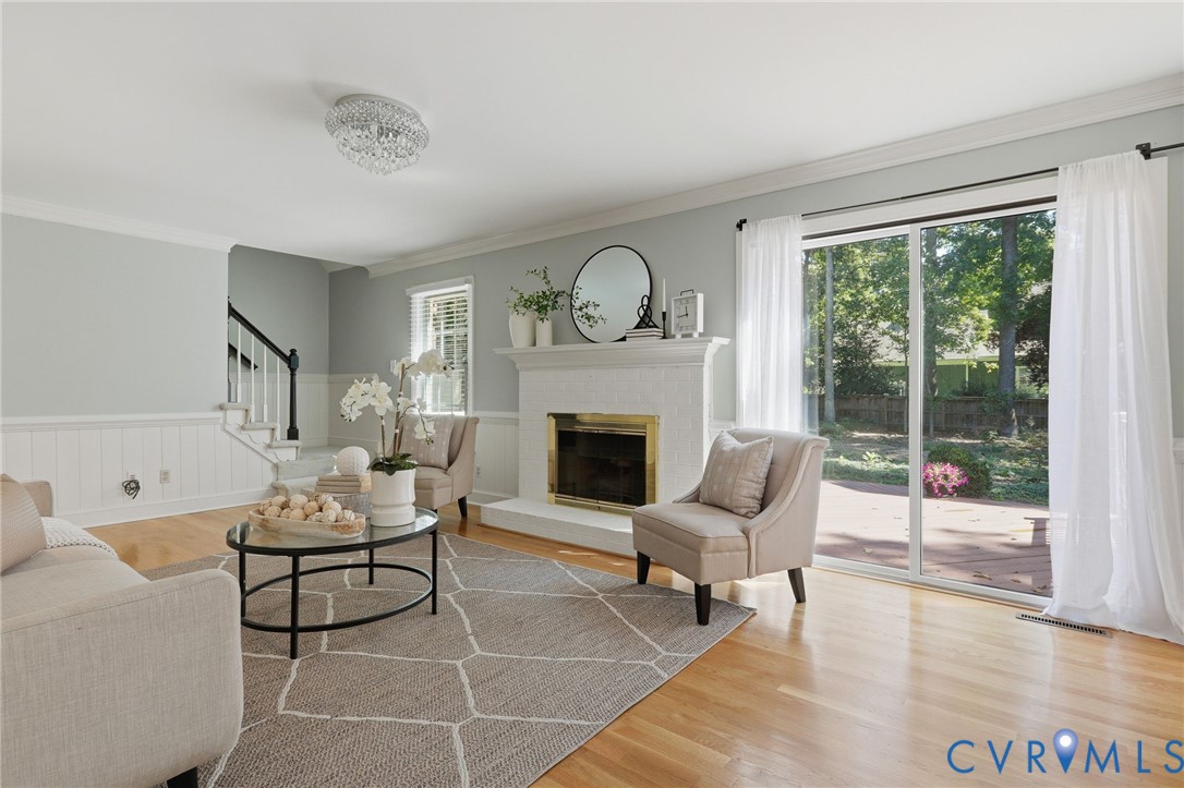 Living area featuring wood-finish flooring, a painted brick fireplace with a mantel, wainscoting, a crystal flush-mount light fixture, and sliding glass doors Living area featuring wood-finish flooring, a painted brick fireplace with a mantel, wainscoting, a crystal flush-mount light fixture, and sliding glass doors