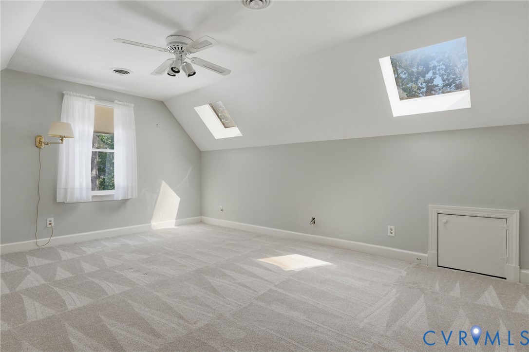 Room featuring sloped ceilings, two skylights, a ceiling fan, wall-mounted lighting, and built-in access panel