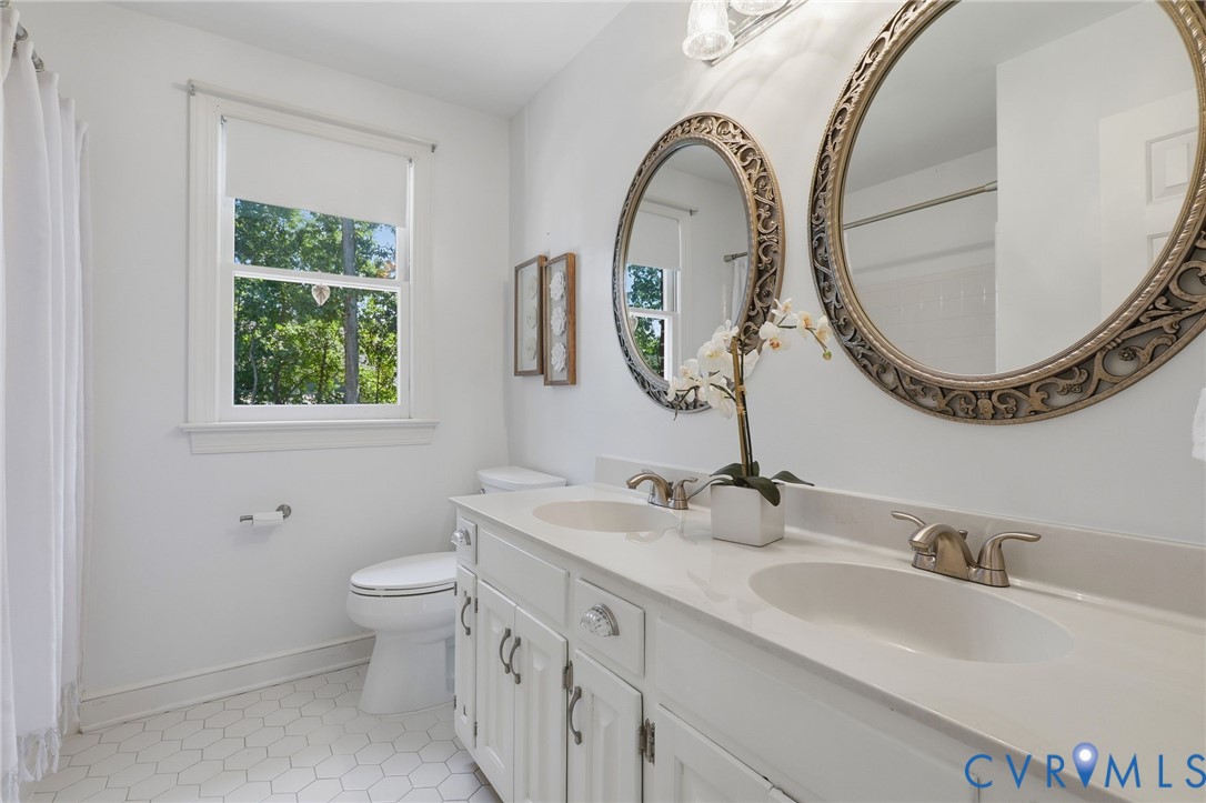 Dual-sink vanity with white cabinetry and integrated stone-top surface, white hexagonal floor tile, and double oval wall-mounted mirrors