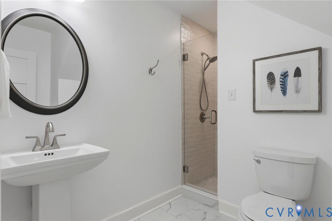 Bathroom featuring a pedestal sink, circular wall-mounted mirror, walk-in shower with a glass door and tiled surround, and stone-finish flooring