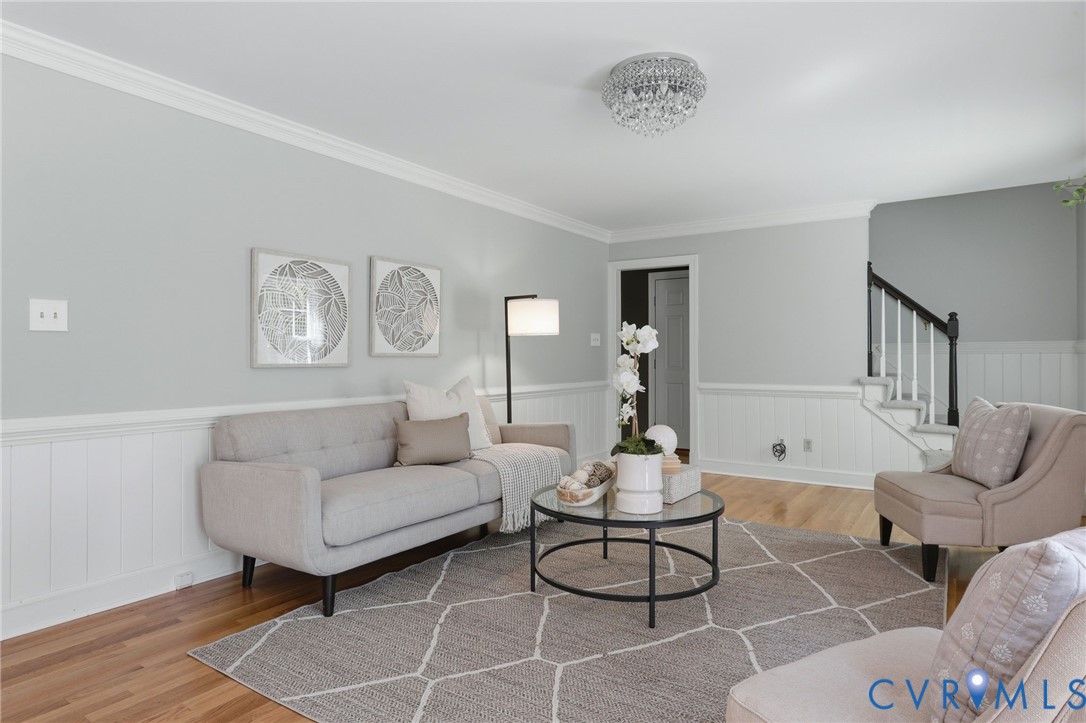 Living area featuring wood-finish flooring, white beadboard wainscoting, crown molding, a flush-mount crystal light fixture, and a staircase with a dark-stained handrail Living area featuring wood-finish flooring, white beadboard wainscoting, crown molding, a flush-mount crystal light fixture, and a staircase with a dark-stained handrail