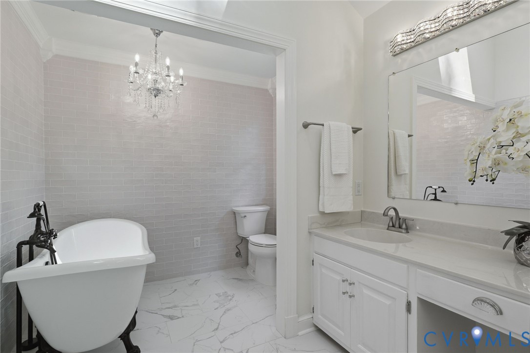 Freestanding soaking tub with a floor-mounted faucet, crystal chandelier, wall-to-wall light-toned subway tile, stone-patterned tile flooring, and a white vanity with integrated lighting