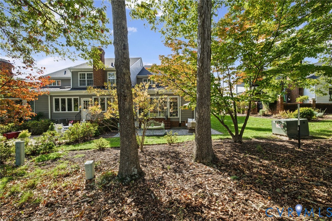 Rear exterior featuring gray horizontal siding, a brick chimney, a stone patio, and mature trees on a lawn