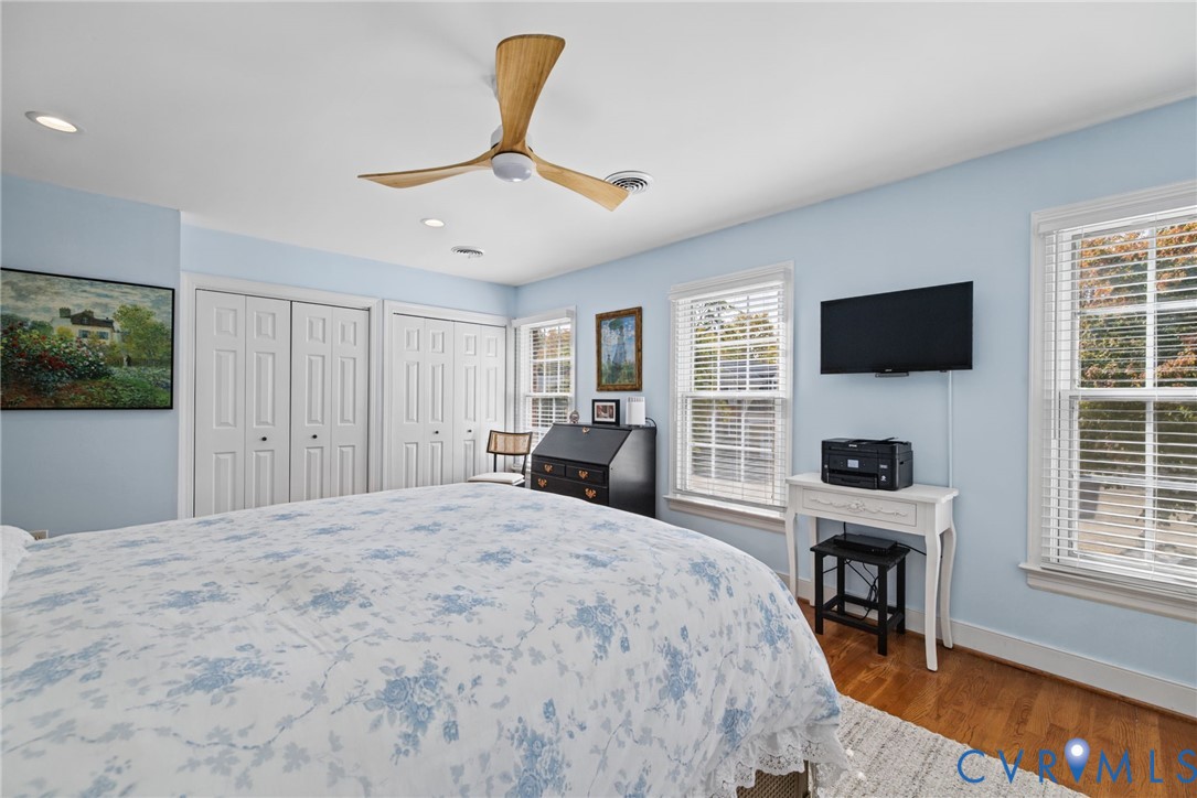 Wood-finish flooring, dual panel-door closets, three-blade ceiling fan, and multi-pane windows with horizontal blinds