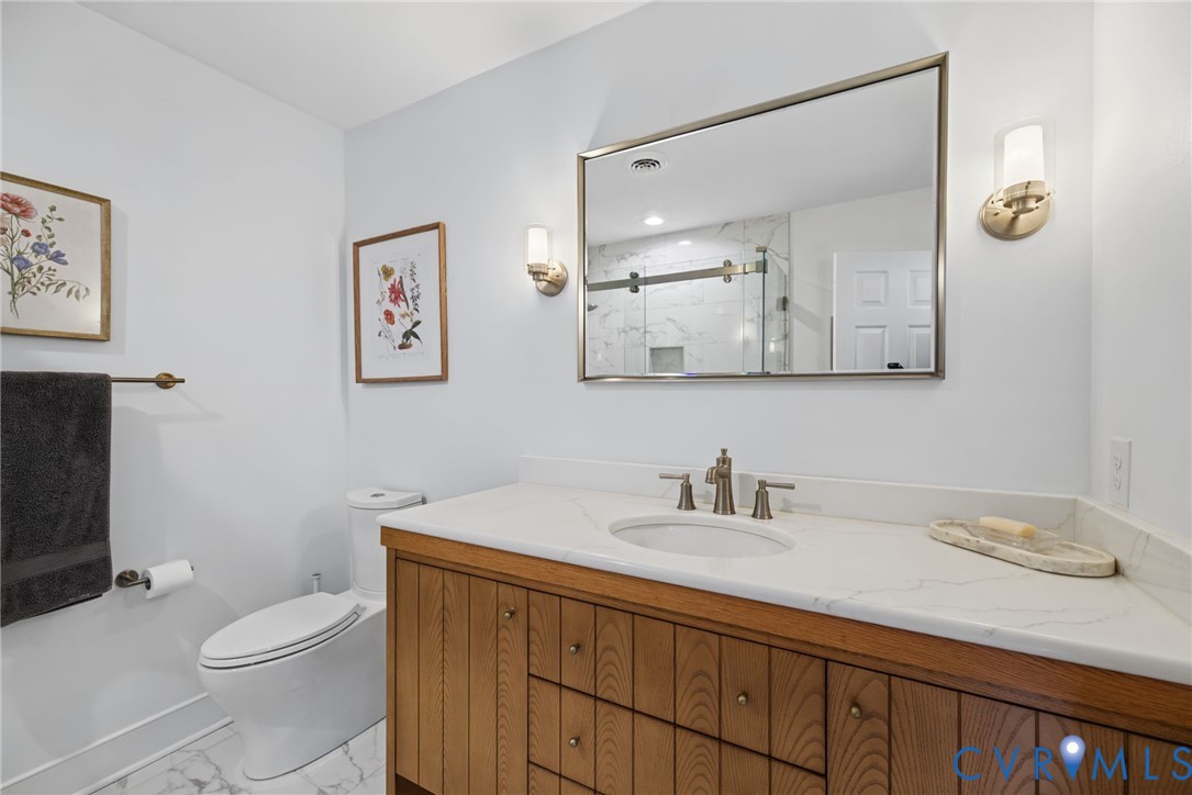 Bathroom featuring a wood-finish vanity with a stone-composite countertop, undermount sink, metallic-finish wall sconces, a framed mirror, and a two-piece elongated toilet