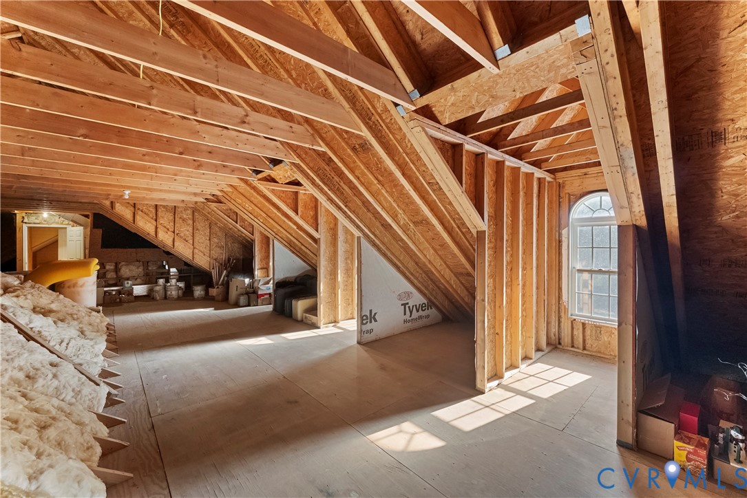 Unfinished attic space featuring exposed wood framing, plywood subflooring, batt insulation, and an arched window