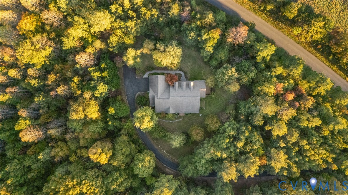 Aerial perspective of a multi-gabled residence situated on a wooded lot, featuring a paved driveway, stone-lined landscaping, and a shingled roof