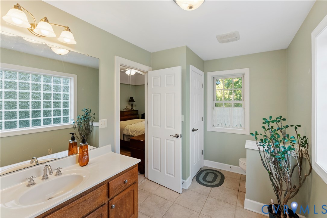 Bathroom featuring a wood-finish vanity with an integrated basin, a wall-to-wall mirror, glass block window, tile flooring, and multi-light fixture