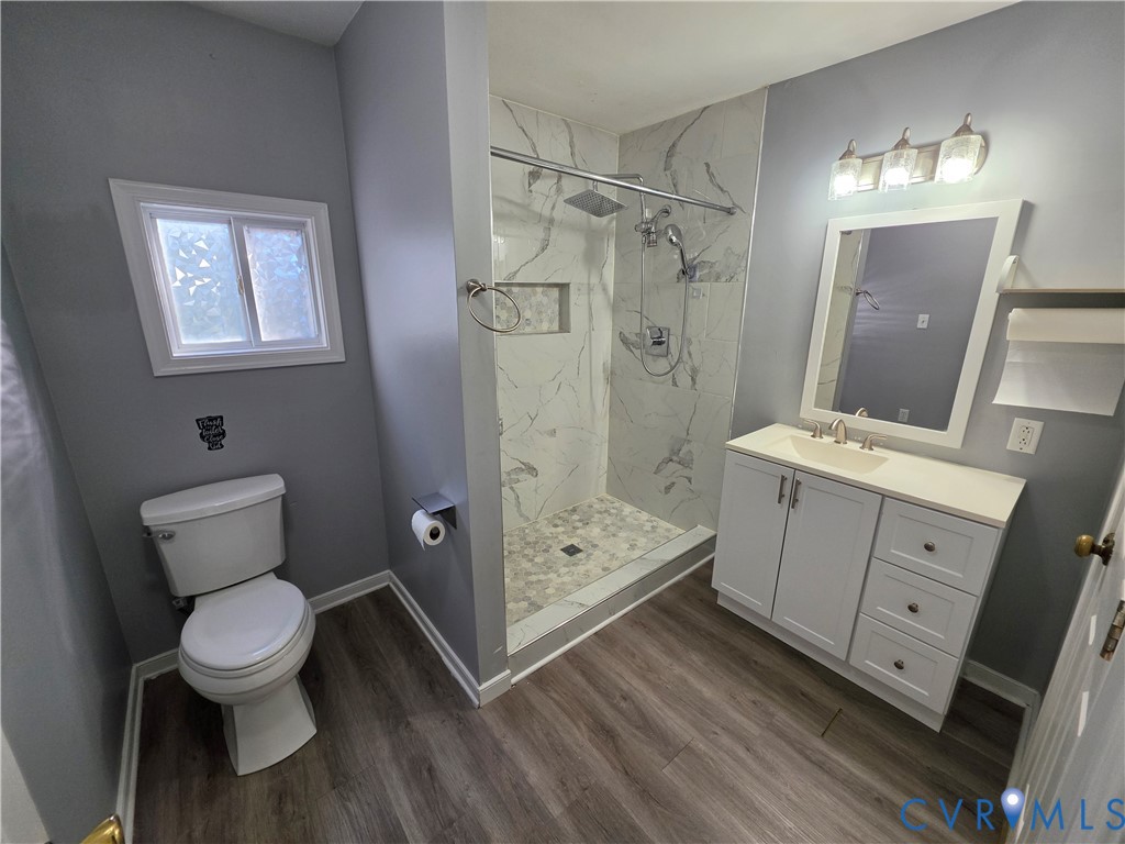 Bathroom featuring a shower stall with stone-patterned tile, built-in shelving niche, overhead rain shower head, wood-finish flooring, and white vanity cabinetry with a solid surface countertop