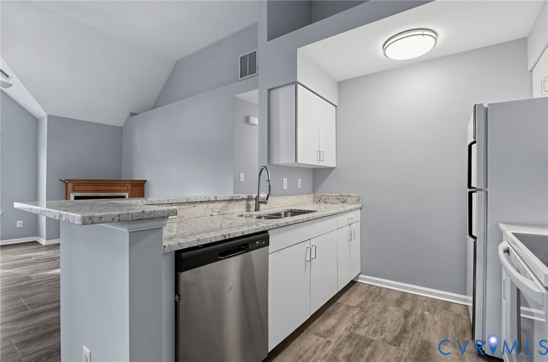 Kitchen featuring a peninsula with stone-finish countertops, stainless steel dishwasher, white cabinetry, wood-finish flooring, and vaulted ceiling