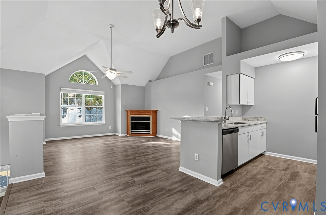 Vaulted ceilings, wood-finish flooring, corner fireplace with wood mantle, and kitchen area featuring white cabinetry with stone-finish countertops