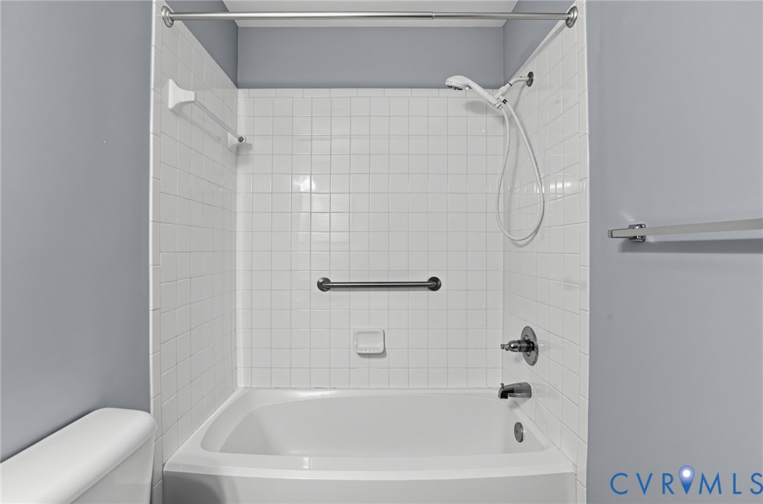Bathtub surround featuring white square ceramic tile, handheld shower fixture, integrated grab bar, and wall-mounted metal towel bars