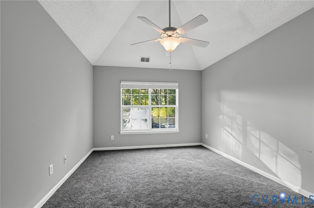 Vaulted ceiling featuring a central multi-blade ceiling fan with integrated light fixture, neutral wall palette, wall-to-wall carpeting, and a double-pane window
