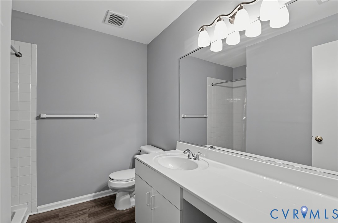 Full-width vanity with integrated sink, expansive vanity mirror, multi-bulb overhead lighting, wood-finish flooring, and tiled shower surround