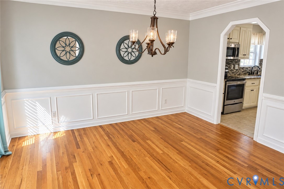 Dining area featuring wood-finish flooring, white picture frame wainscoting, an arched doorway, and a multi-arm metal chandelier Dining area featuring wood-finish flooring, white picture frame wainscoting, an arched doorway, and a multi-arm metal chandelier
