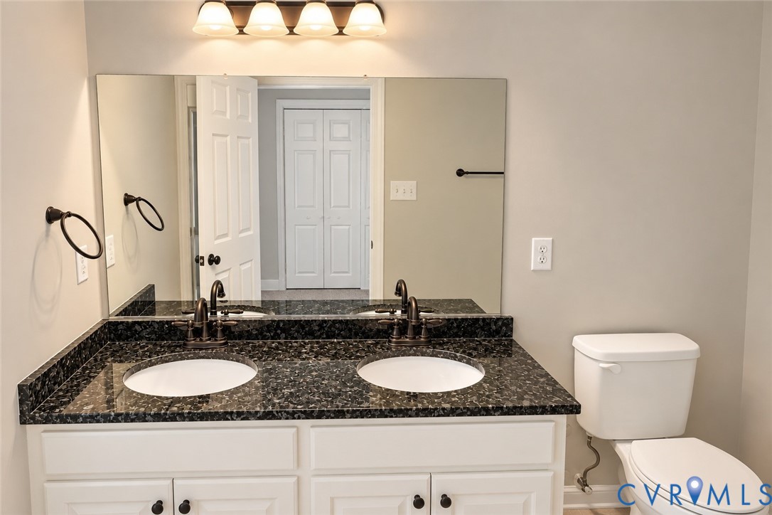 Dual-sink vanity with a stone countertop, white cabinetry, polished metal fixtures, and a wall-mounted light fixture