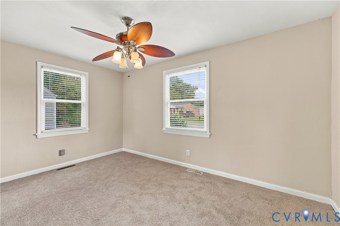 Neutral-toned interior featuring carpeting, a multi-blade ceiling fan with integrated lighting, and dual windows with horizontal blinds