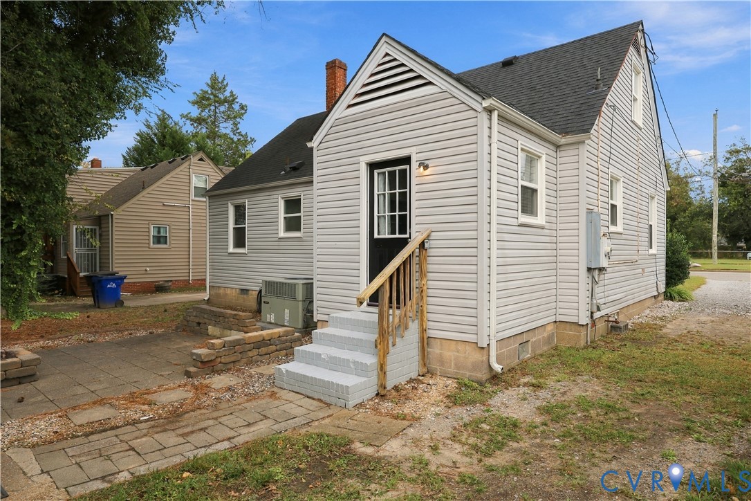 Light gray vinyl siding, gabled roof, paved stone patio, retaining wall, and exterior wooden stair railing