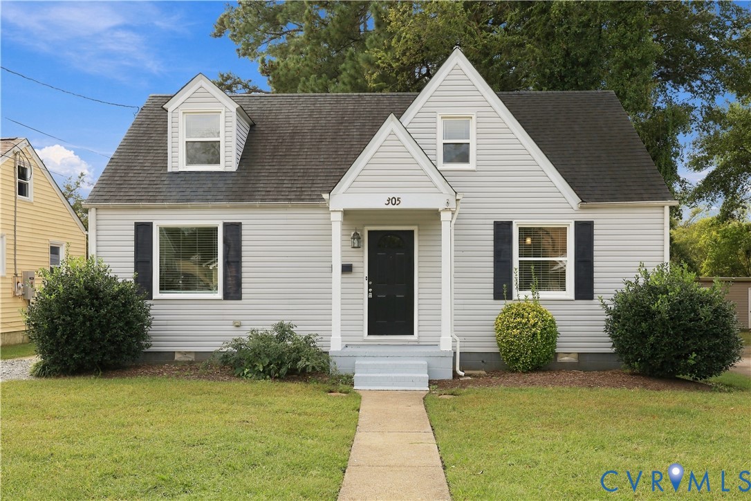 Cape Cod exterior featuring light gray horizontal siding, dark window shutters, a covered front entryway with columns, and a gabled roofline with dual dormers