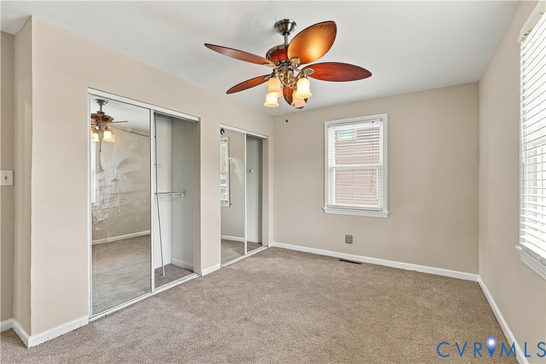 Room featuring neutral-toned carpeting, dual sliding mirror-paneled closet doors, a multi-blade ceiling fan with integrated lighting, and white horizontal window blinds