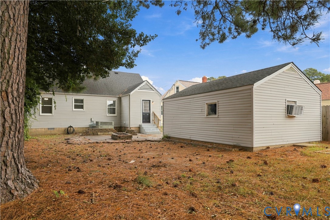 Exterior featuring light-gray horizontal siding, a detached garage, a stone fire pit, and mature pine trees
