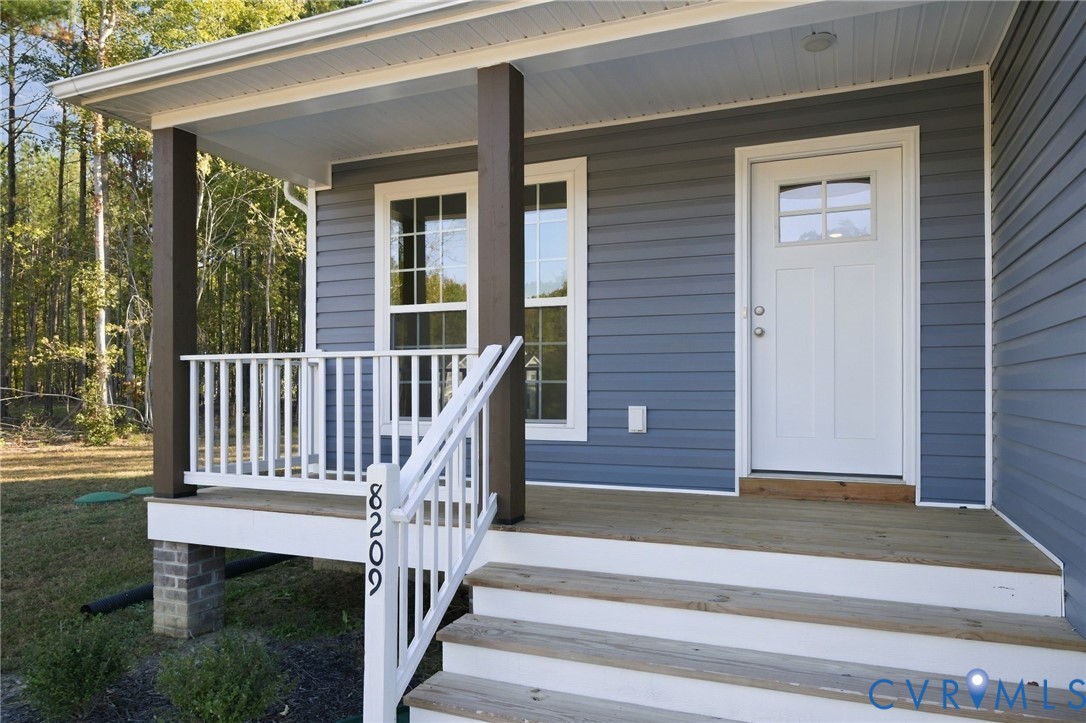 Covered front porch featuring wood-finish decking, white railings, dark-tone support posts, and a white six-panel door with glass inlay