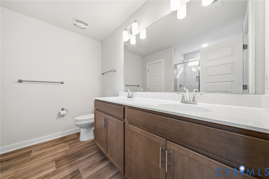 Dual-sink vanity featuring a white countertop, wood-finish cabinetry, full-width mirror, and multi-bulb light fixture, accompanied by wood-finish flooring