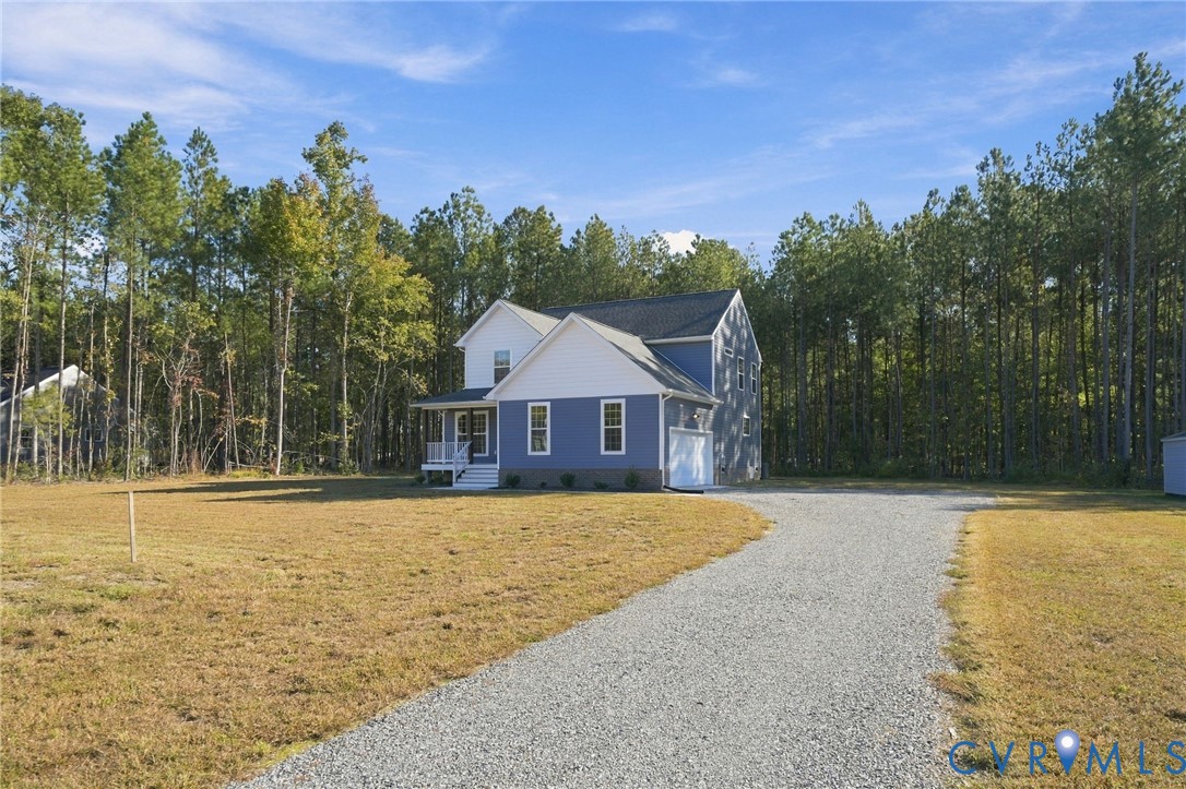 Two-story residence featuring blue siding with white trim, a covered front porch, attached garage, and a gravel driveway situated on a cleared lot