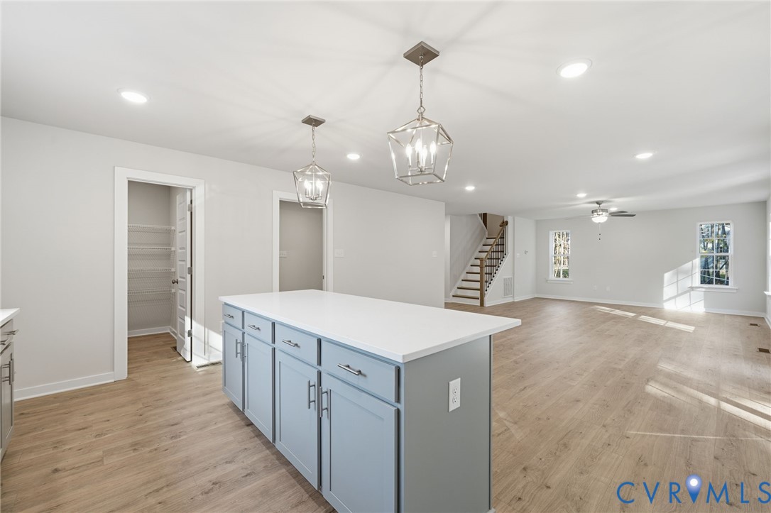 Kitchen island featuring light-toned cabinetry and a solid-surface countertop, wood-finish flooring, recessed lighting, geometric pendant fixtures, and a walk-in pantry with wire shelving