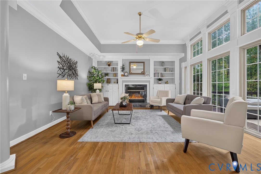 Living room featuring wood-finish flooring, floor-to-ceiling windows, crown molding, built-in shelving, and a fireplace with stone surround Living room featuring wood-finish flooring, floor-to-ceiling windows, crown molding, built-in shelving, and a fireplace with stone surround