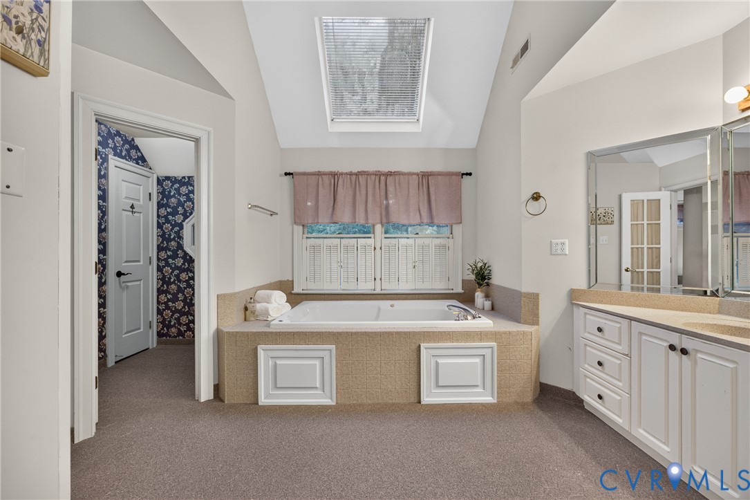 Built-in soaking tub with a textured surround and recessed cabinet panels, a vaulted ceiling featuring a skylight, and white cabinetry with a stone-finish countertop