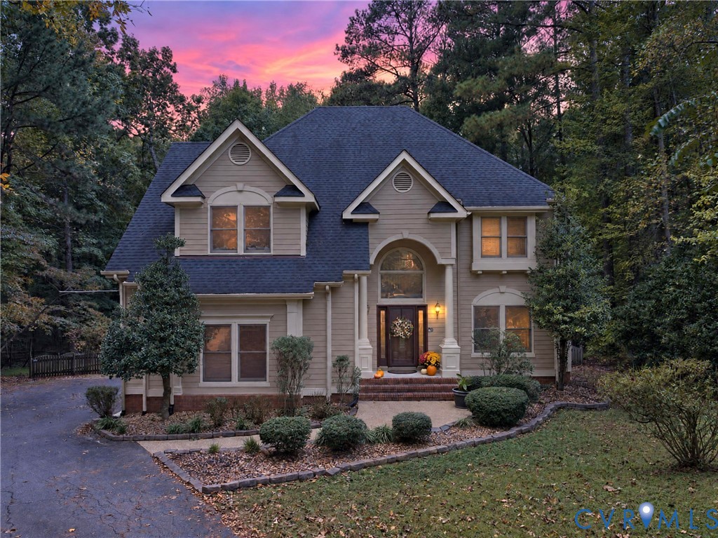 Two-story exterior featuring tan horizontal siding, an arched entryway with column accents, a multi-gabled roofline, and landscaped brick-bordered garden beds Two-story exterior featuring tan horizontal siding, an arched entryway with column accents, a multi-gabled roofline, and landscaped brick-bordered garden beds