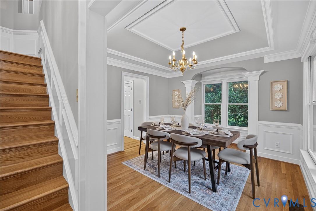 Dining area featuring a tray ceiling with crown molding, wainscoting, wood-finish flooring, a multi-arm chandelier, and a wood-finish staircase with white handrails