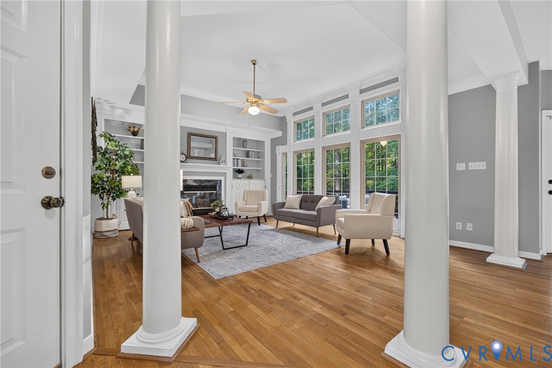 Living area featuring wood-finish flooring, floor-to-ceiling windows, dual decorative columns, and built-in shelving flanking a fireplace Living area featuring wood-finish flooring, floor-to-ceiling windows, dual decorative columns, and built-in shelving flanking a fireplace