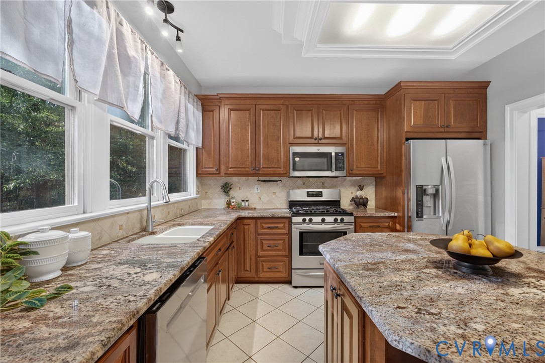 Kitchen featuring wood cabinetry, natural stone countertops, tile backsplash, stainless steel appliances, and tile flooring