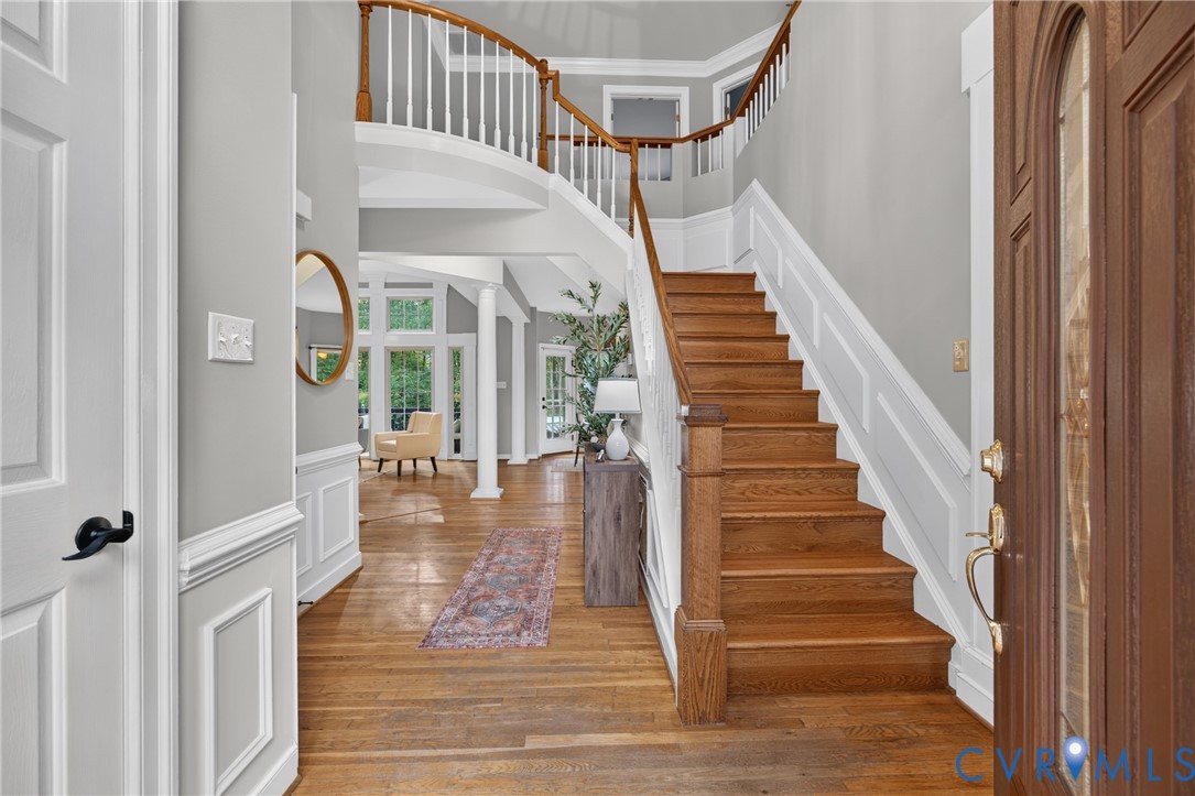Grand foyer featuring a wood-finish staircase with decorative balusters, wood-finish flooring, wainscoting, and a curved landing Grand foyer featuring a wood-finish staircase with decorative balusters, wood-finish flooring, wainscoting, and a curved landing