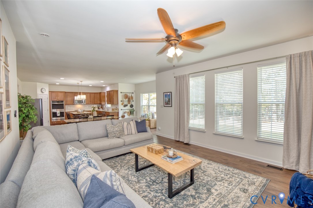 Open-concept interior featuring wood-finish flooring, a multi-blade ceiling fan with integrated lighting, and a kitchen equipped with built-in stainless steel appliances and wood cabinetry