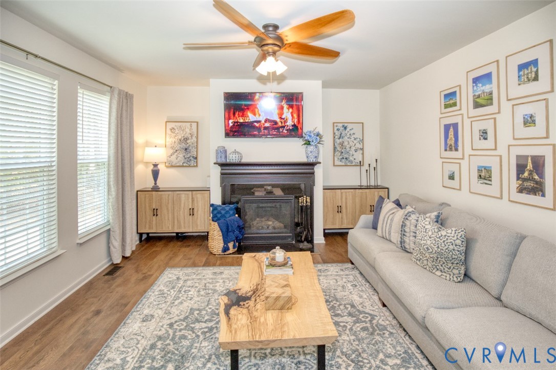 Living area featuring wood-finish flooring, a fireplace with a black surround and mantel, built-in cabinetry, and a ceiling fan