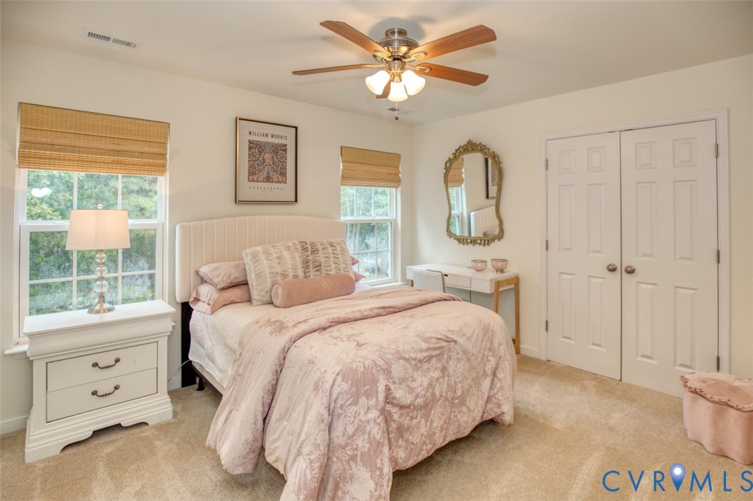 Neutral carpeted flooring, dual-window configuration, wood-finish ceiling fan with integrated lighting, and white paneled double-door closet