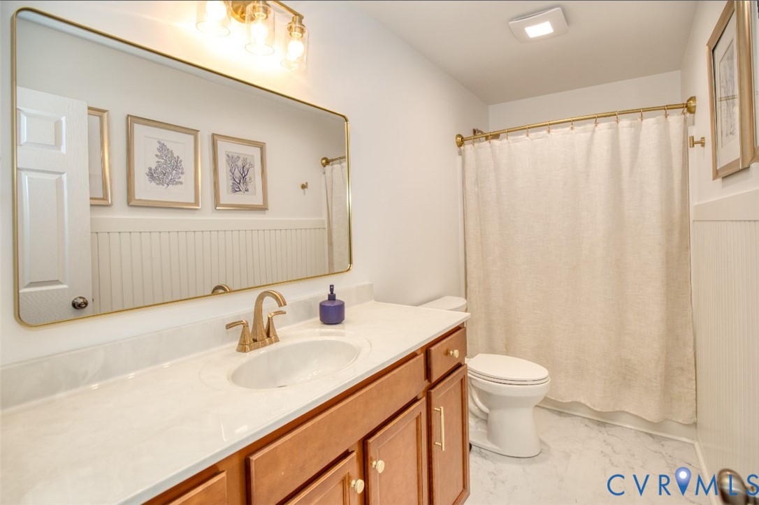 Bathroom featuring a vanity with a light-toned countertop, beadboard wainscoting, a large brass-framed vanity mirror, brass-finish faucet hardware, and a matching brass shower curtain rod
