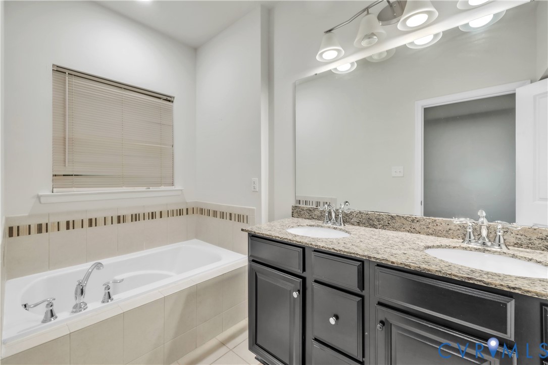 Dual-sink vanity with dark cabinetry, stone-finish countertop, soaking tub with tile surround, and expansive wall-mounted vanity mirror