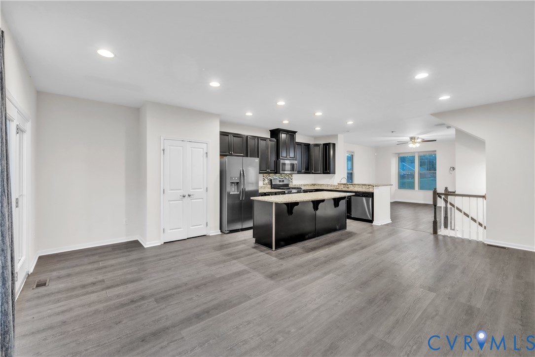 Open-concept kitchen featuring dark cabinetry, a central island with a stone-finish countertop, stainless steel appliances, recessed lighting, and wood-finish flooring