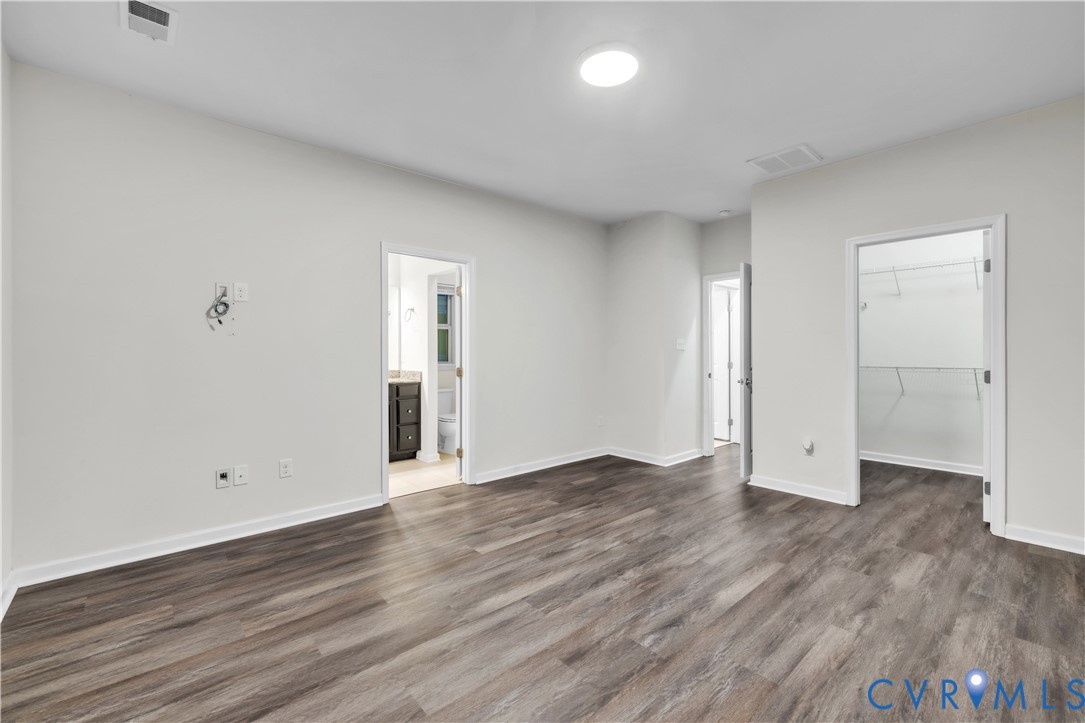 Neutral-toned interior space featuring wood-finish flooring, flush-mount ceiling lighting, and two interior doorways leading to an en-suite area and a walk-in closet with built-in shelving
