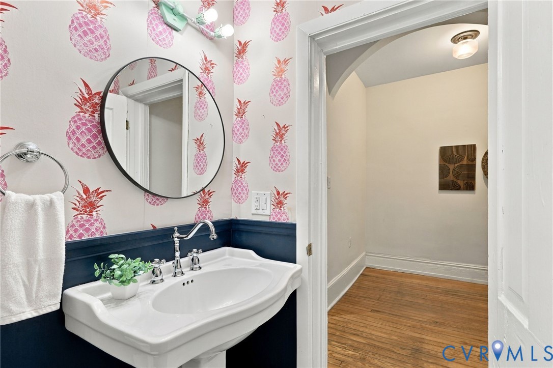 Wall-mounted pedestal sink with chrome faucet, circular wall mirror, custom patterned wallpaper, and navy blue wainscoting
