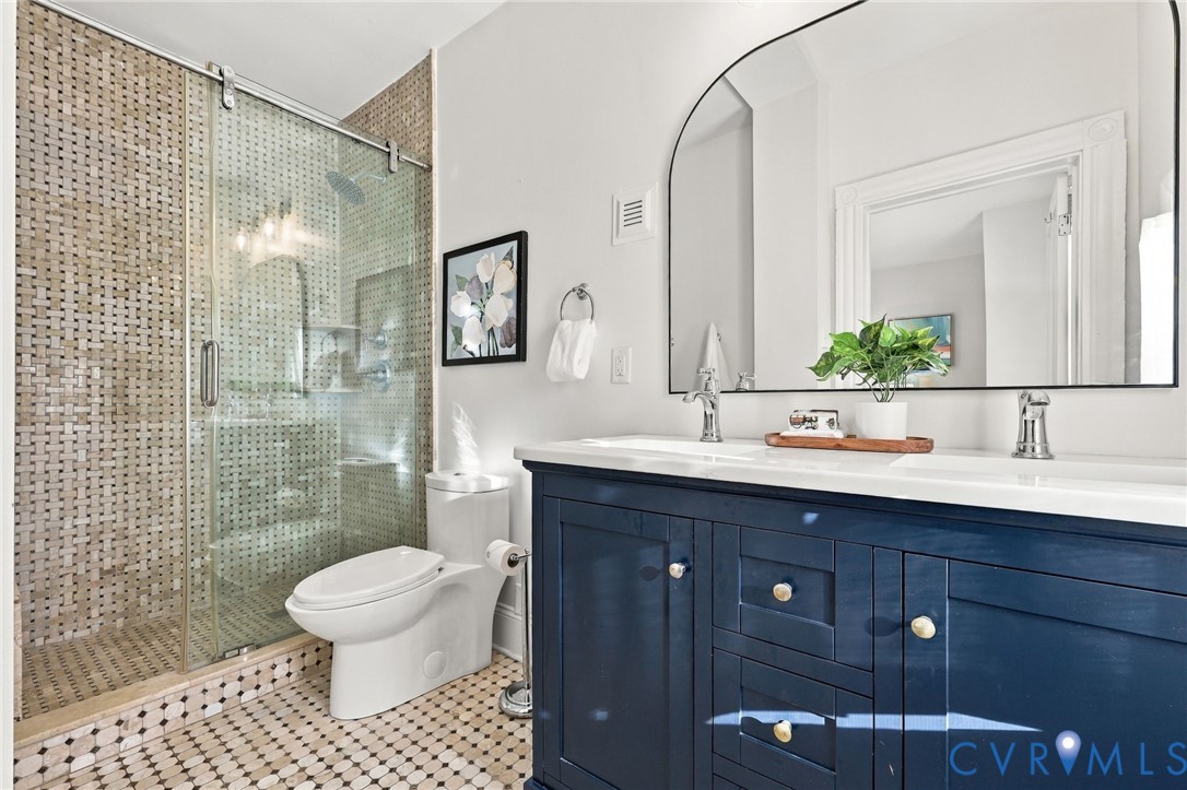 Bathroom featuring a double-sink vanity with a solid-surface countertop, an arched wall-mounted mirror, and a walk-in shower with a sliding glass door and mosaic tile surround