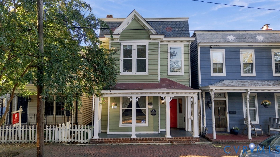 Historic architectural exterior featuring sage-colored siding, a covered front porch with decorative gingerbread trim, a brick-foundation stoop, and a gabled roofline