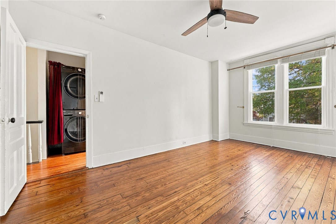 Front bedroom #2featuring wood-finish flooring, a ceiling-mounted fan with light fixture, double-pane windows, and a laundry area equipped with a stacked washer and dryer set