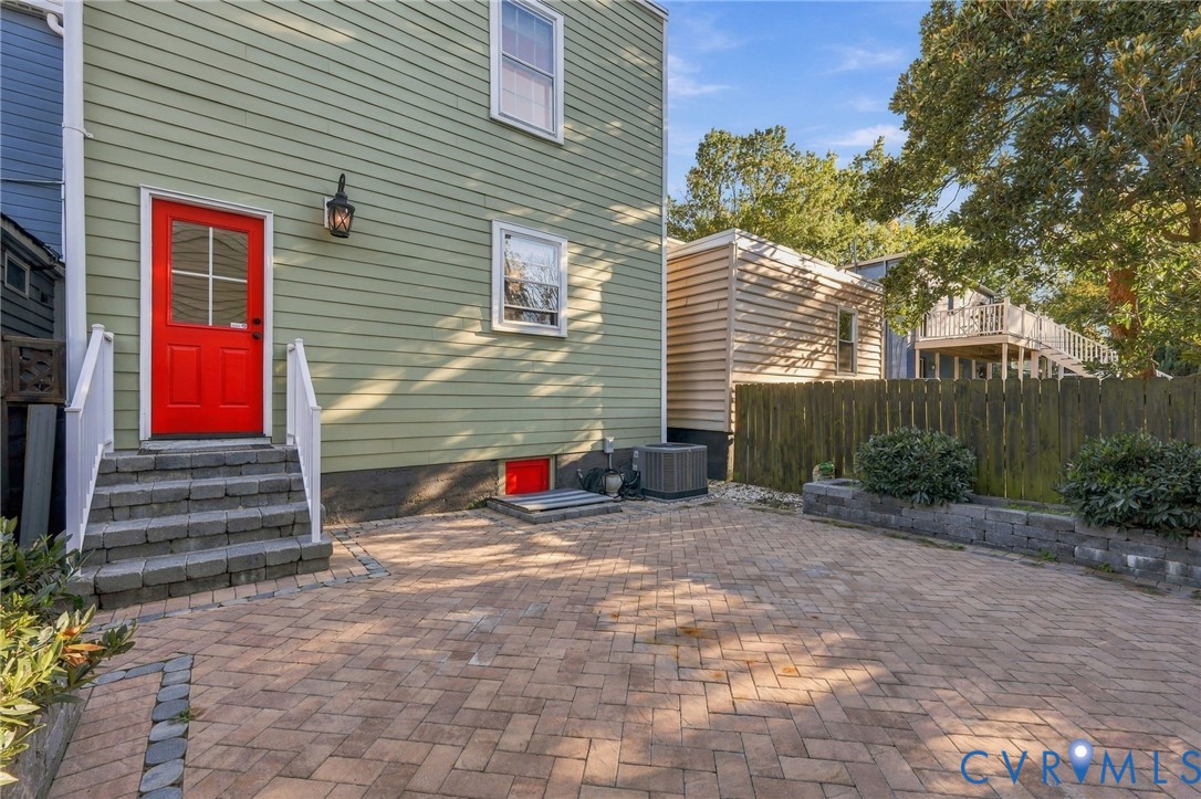 Exterior featuring horizontal lap siding, a paved brick patio, stone-block steps, a red entry door, and a wood privacy fence