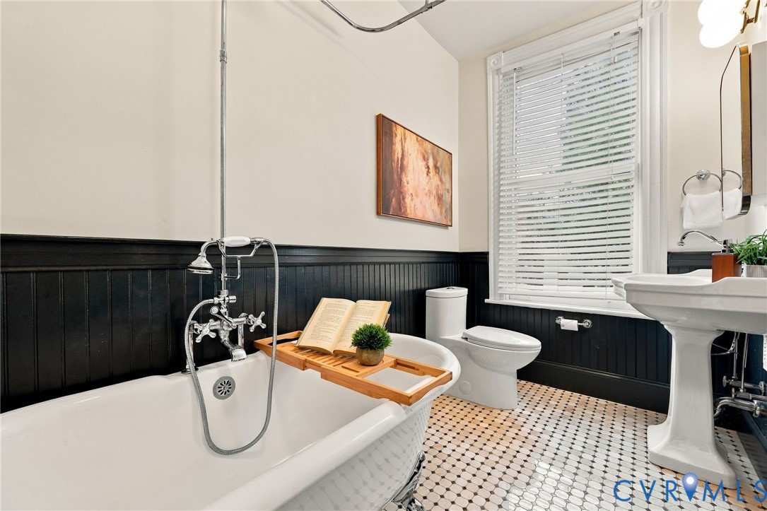 Hall full bath featuring lcawfoot soaking tub with exposed chrome plumbing fixtures, black beadboard wainscoting, pedestal sink, and geometric tile flooring