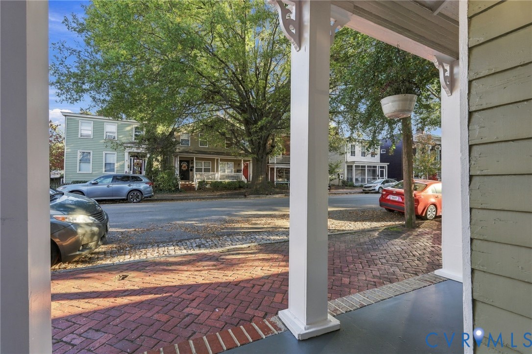 View of the street from the covered front porch featuring white support columns, horizontal lap siding, and a red brick herringbone walkway